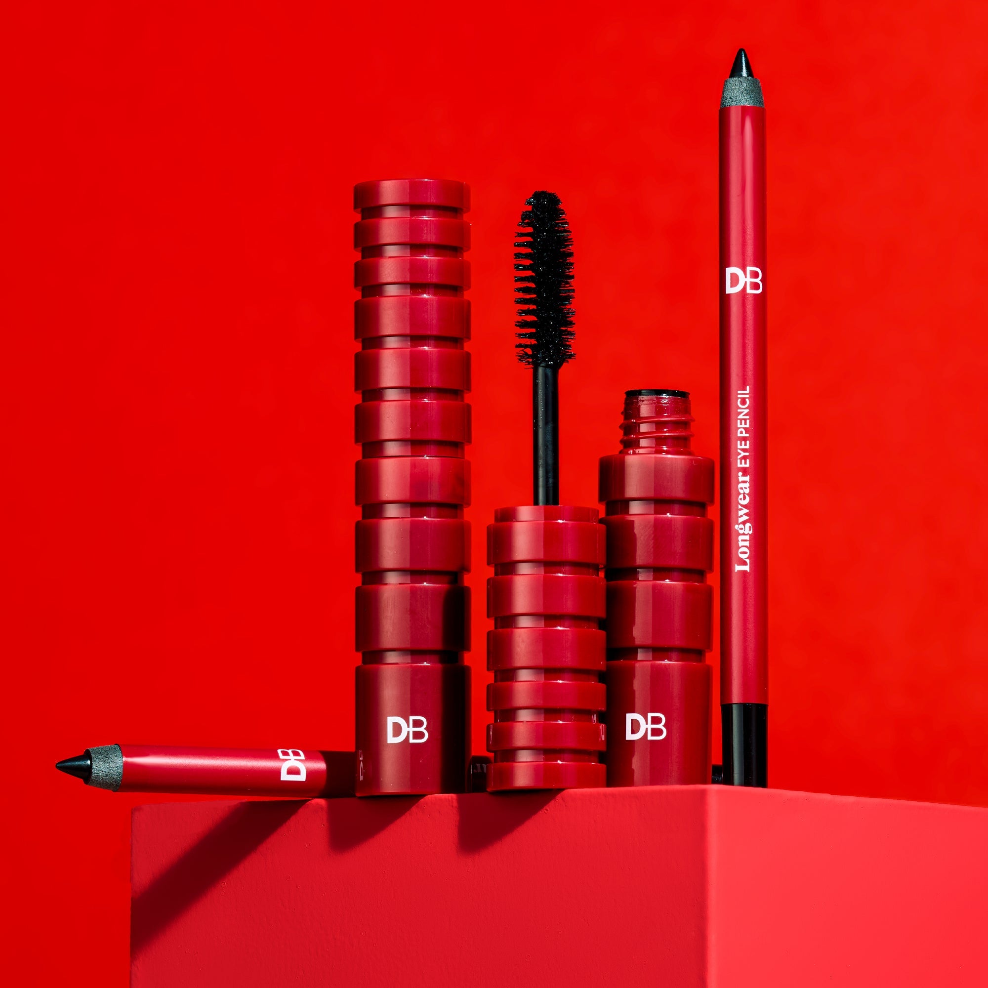 Makeup products including a mascara, eyeliner pencil, and other items on a red background with 'DB' branding.