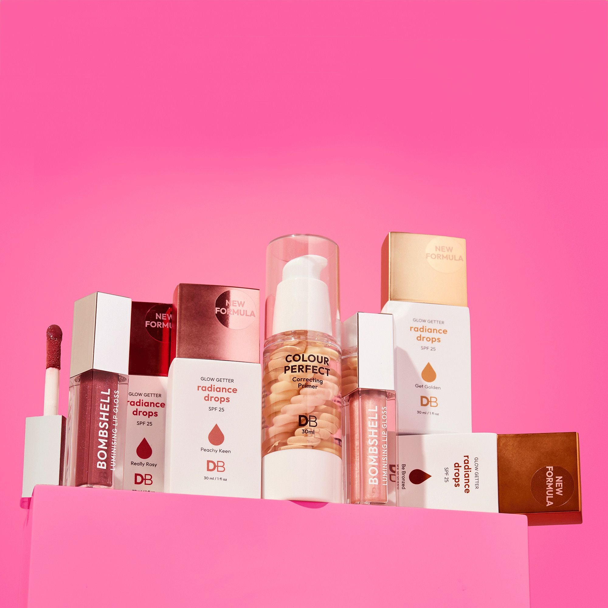 A collection of luxury makeup products on a pink background, featuring lipsticks, radiance drops, and a foundation bottle, elegantly arranged.