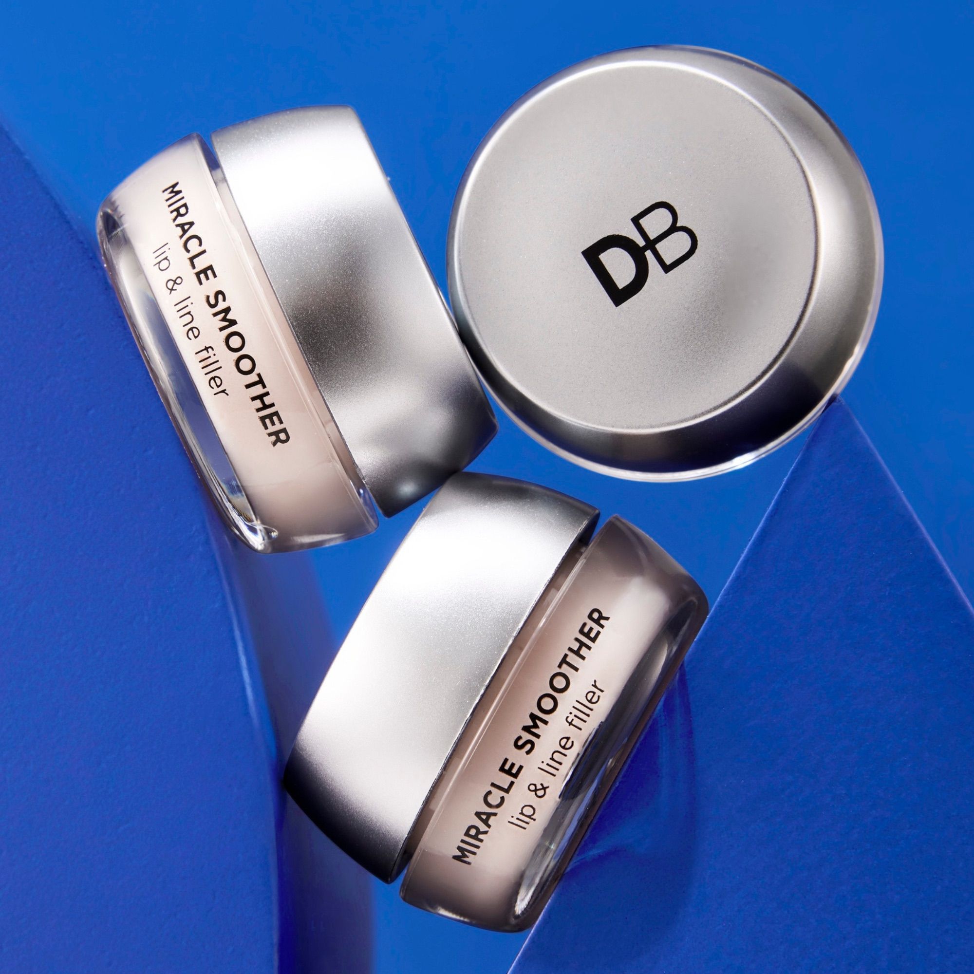 Three silver jars of "Miracle Smoother Lip & Line Filler" with "DB" logo on a blue background, showcasing luxury skincare design.