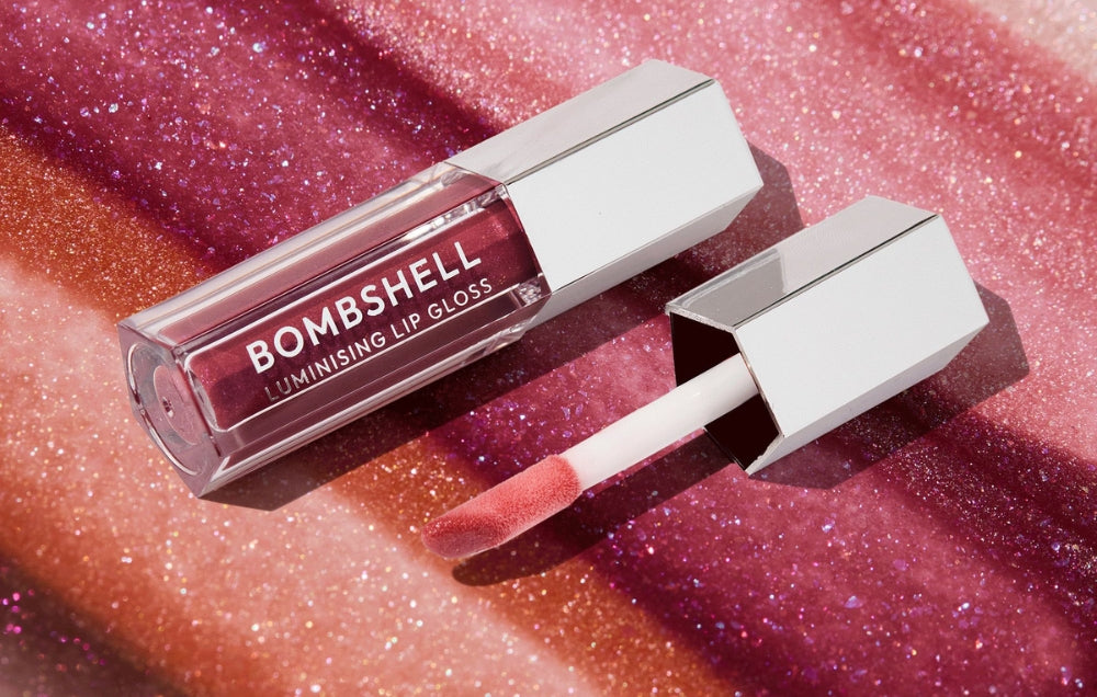 Bombshell Luminising Lip Gloss in sleek silver packaging against a glittery pink backdrop, with applicator showing glossy pink hue.