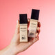Two Designer Brands makeup bottles—Rise & Prime Luminescent Primer and Luminous Hydrating Foundation—held against a pink background.